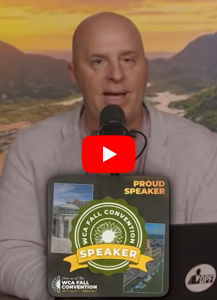 Bald man speaks into a microphone at a convention, wearing a light blazer and a badge that reads 'WCA Fall Convention Speaker.'